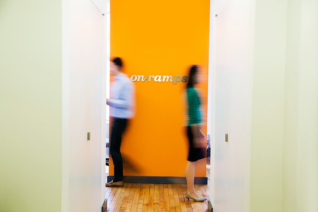 People walking past the On-Ramps logo hanging on an orange wall in the New York City office