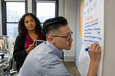 An On-Ramper writes on a giant sticky note while a second On-Ramper looks on.